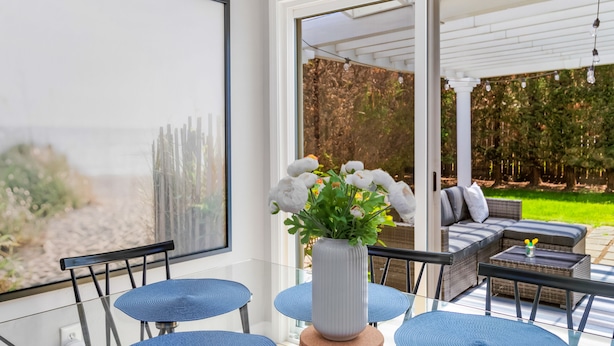 Een moderne patio met een glazen tafel, blauwe stoelen, een witte vaas met bloemen en uitzicht op een groene tuin door een glazen deur.