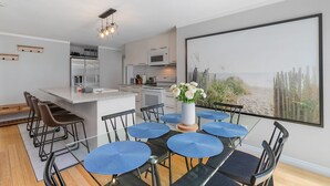 Een moderne keuken met witte apparaten, een eethoek met blauwe stoelen en een centraal keukenblok, en een groot raam met uitzicht op het strand.