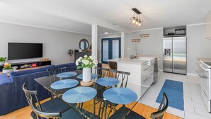 Een moderne keuken met een koelkast, fornuis en spoelbak. Er is een eethoek met een tafel en stoelen, en een zithoek met een bank en een televisie.
