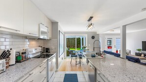 Een moderne keuken met witte kasten, granieten aanrechtbladen en een centraal eiland. Er is een zithoek met een blauwe bank en uitzicht op de omgeving.