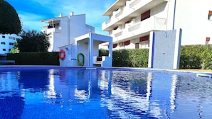 A swimming pool with a clear blue water surface, surrounded by modern buildings with balconies and a hedge. - Vilamoura Twins 1 With Pool by Homing (Loulé)