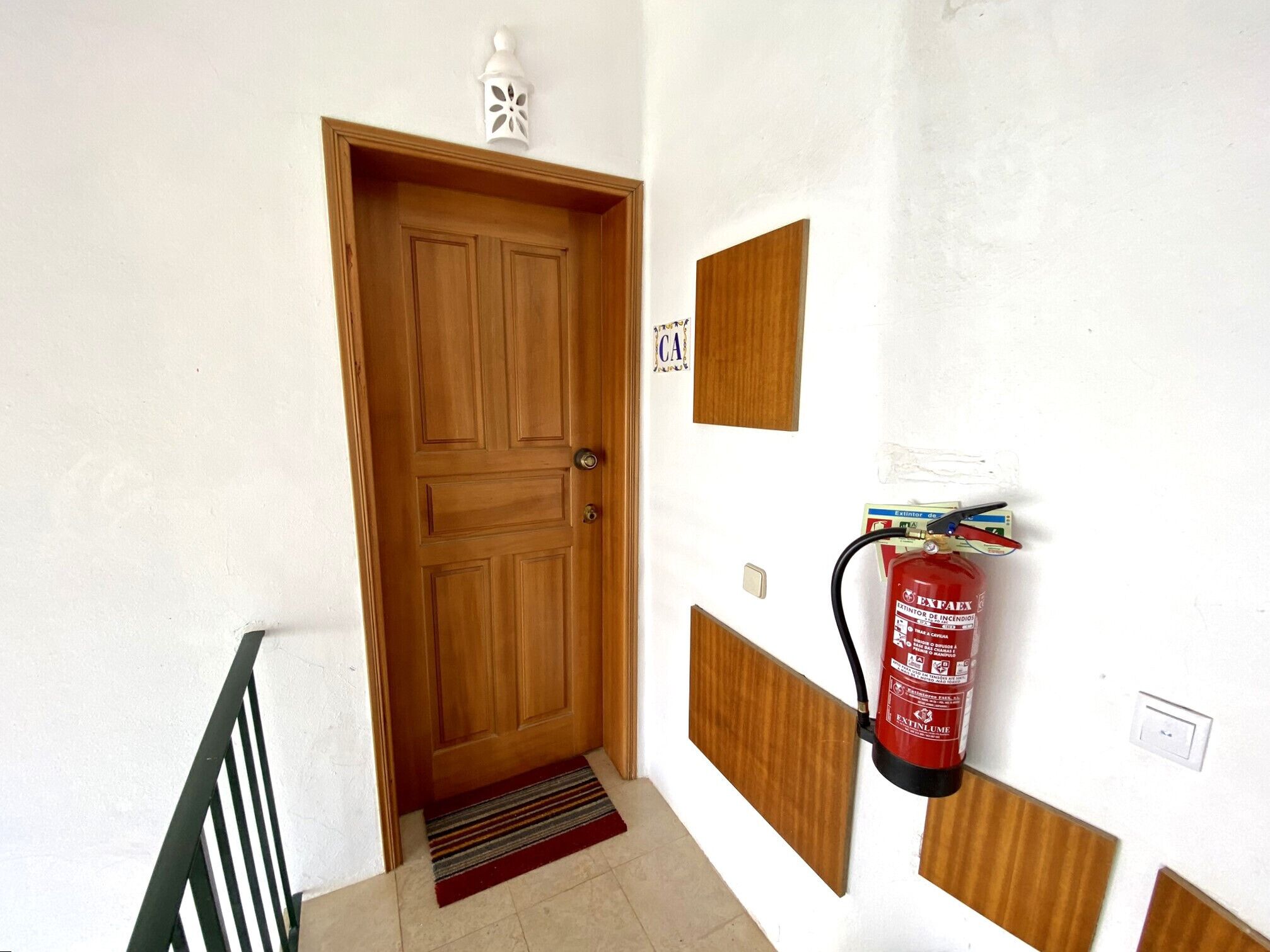 Eine Holztür mit Fußmatte, ein an der Wand befestigter Feuerlöscher und ein kleiner, weißer Laternenleuchter darüber.