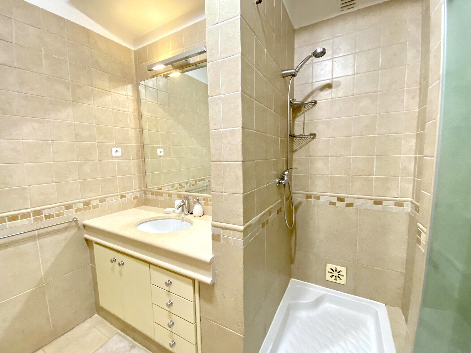 Une salle de bain avec des carreaux de couleur beige, un lavabo à deux cuves, un miroir, une douche avec un pommeau de douche amovible et des toilettes.
