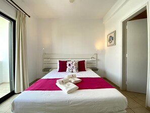 Hotellihuone, jossa on sänky, ikkuna verhoilla, yöpöytä lampun kanssa ja ovi.