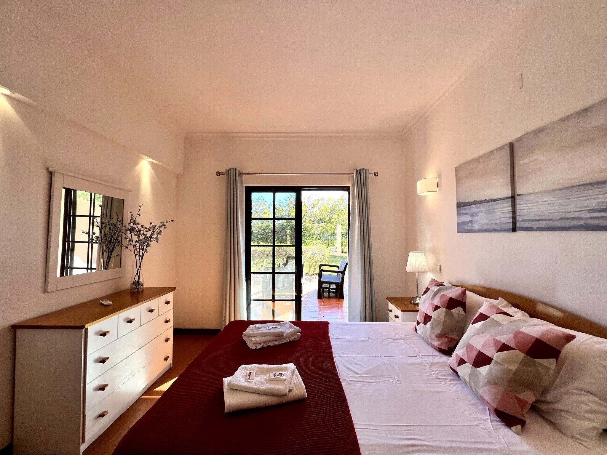 Habitación de hotel con cama, cómoda, espejo, ventana con cortinas, una silla y un cuadro en la pared.