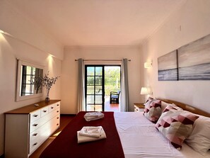 A hotel room with a bed, a dresser, a mirror, a window with curtains, a chair, and a painting on the wall. - Vilamoura Paradise Golf With Pool (Loulé)