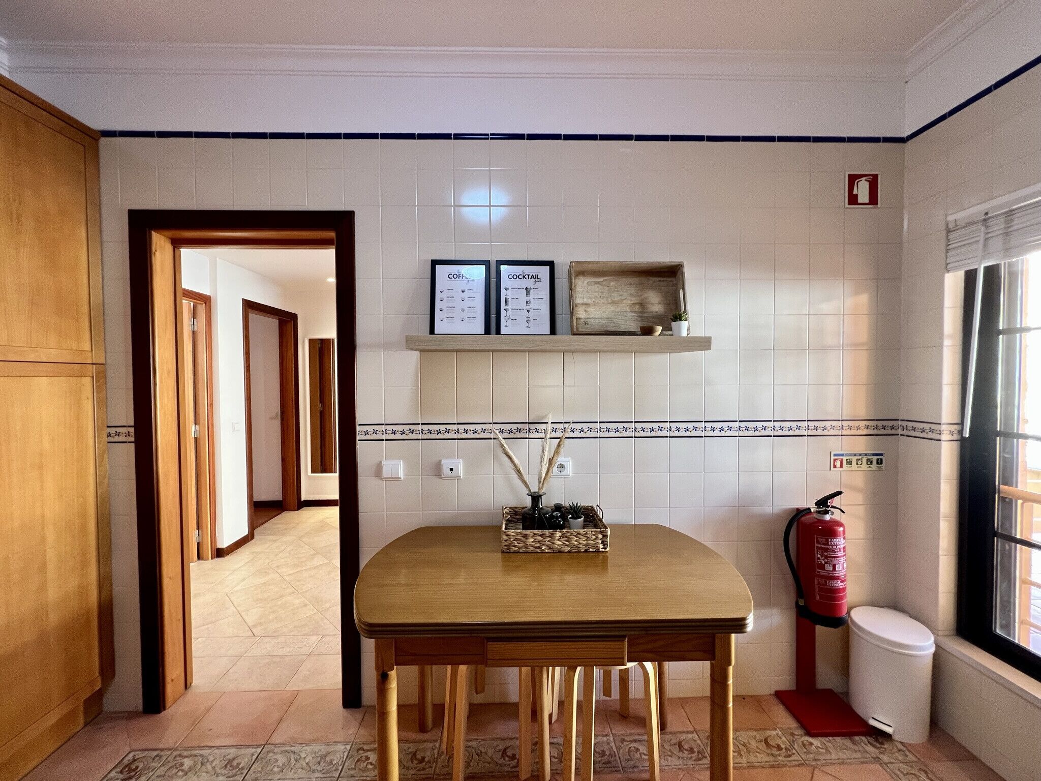 A kitchen with a wooden table, a shelf with framed menus, a fire extinguisher, and a window.