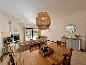 A modern living room with a wooden dining table, sofa, and a large window with a view of greenery. - Vilamoura Paradise Golf With Pool (Loulé)