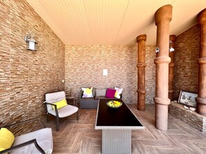 Ein Zimmer mit einer Steinwand, einer Holzbalkendecke, einem schwarzen Tisch mit einer Vase, einem grauen Sofa mit bunten Kissen und einem Stuhl mit einem gelben Kissen.