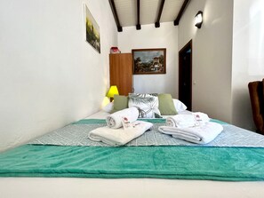 Ein ordentlich gemachtes Bett mit zwei gerollten Handtüchern, einer grünen Decke und einem gemusterten Bettbezug. An der Wand hängt ein Gemälde und auf einem Nachttisch steht eine Lampe.