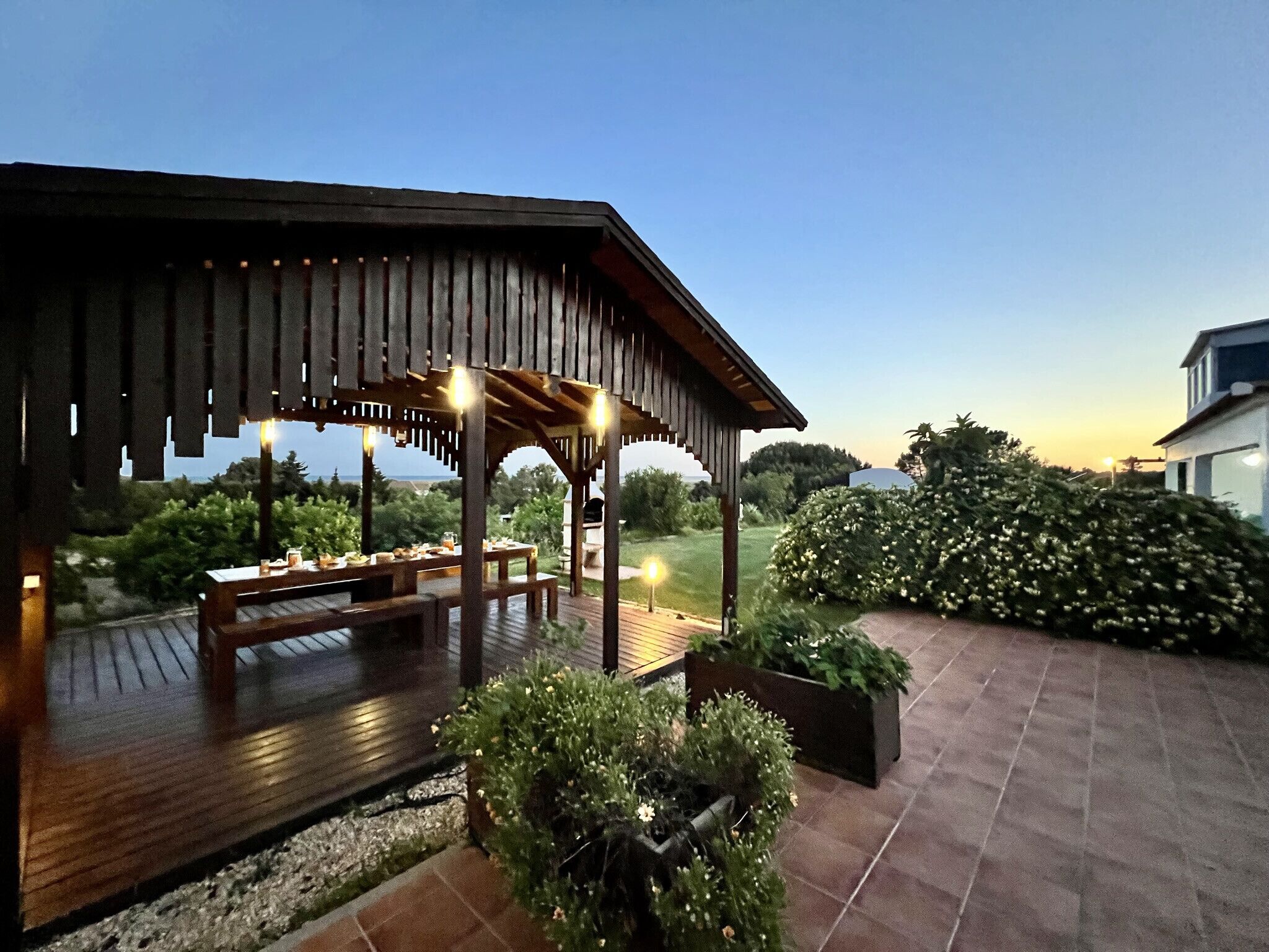 Un espace extérieur abrité, aménagé pour les repas, avec des bancs et des tables en bois, entouré de plantes en pot et offrant une vue sur le paysage.