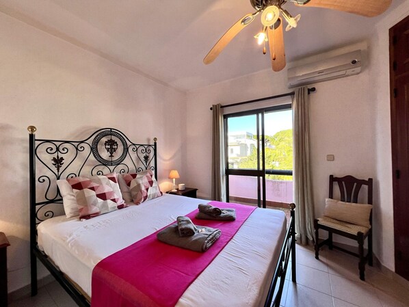 A neatly made bed with a pink bedspread, a ceiling fan, and a window with curtains.