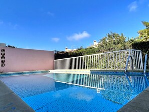 A swimming pool with a white fence, clear blue water, and a view of buildings and trees in the background.