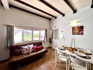 Ein modernes Wohnzimmer mit einem braunen Ledersofa, einem Esstisch, der für ein Frühstück gedeckt ist, und einem großen Fenster mit Vorhängen.