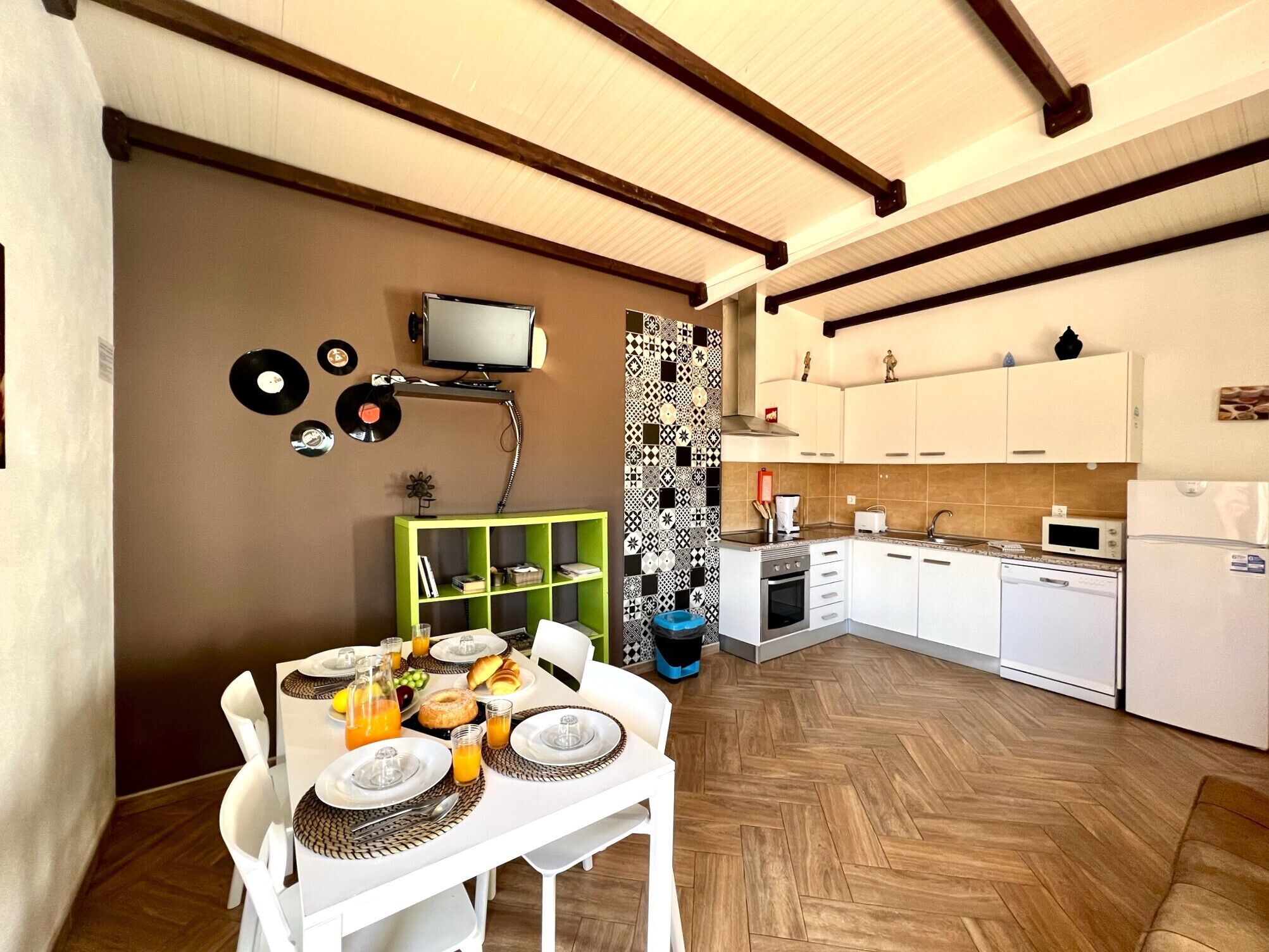 A compact kitchen with white cabinets, a microwave, and a dishwasher. There is a dining table with plates and glasses, and a television mounted on the wall.
