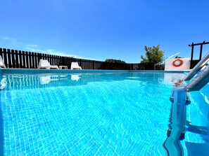 Ein klares, blaues Schwimmbecken mit einer gefliesten Liegefläche, weißen Liegestühlen und einem Holzzaun im Hintergrund.