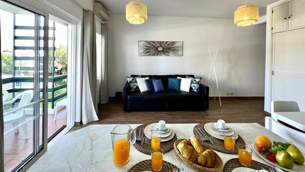 A modern living room with a dark-colored sofa, a dining table set with breakfast, and a balcony with outdoor furniture. - Vilamoura Central 4 With Pool by Homing (Loulé)