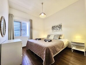 Ein Schlafzimmer mit einem Bett, einem Nachttisch, einer Lampe, einem Fenster mit Vorhängen und einem an der Wand hängenden Kunstwerk.