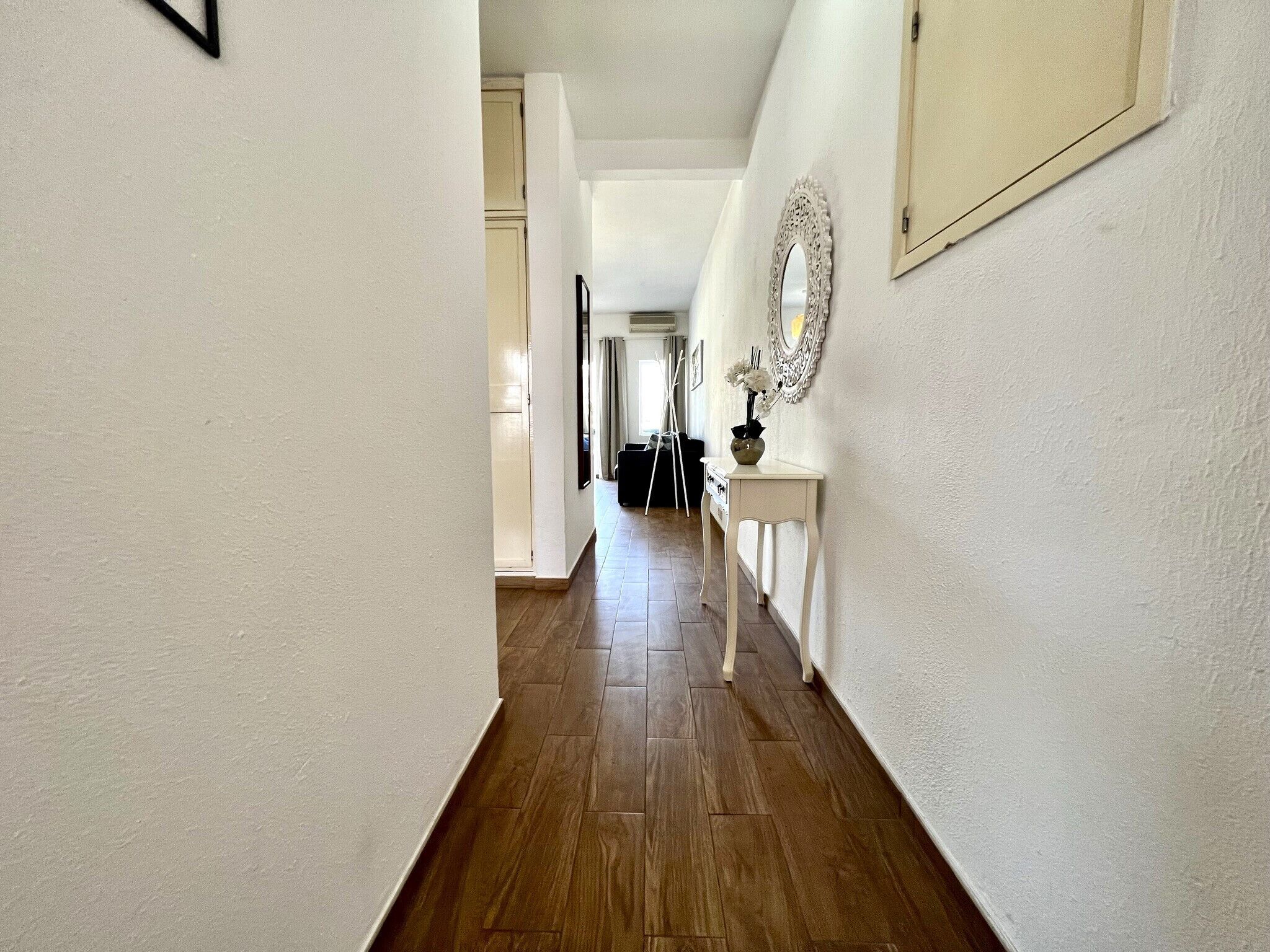 Un pasillo con suelo de madera, paredes blancas y una consola blanca con espejo y jarra.