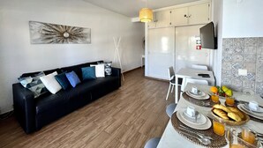 A modern living room with a black sofa, a dining table set with plates, glasses, and a fruit basket. - Vilamoura Central 4 With Pool by Homing (Loulé)