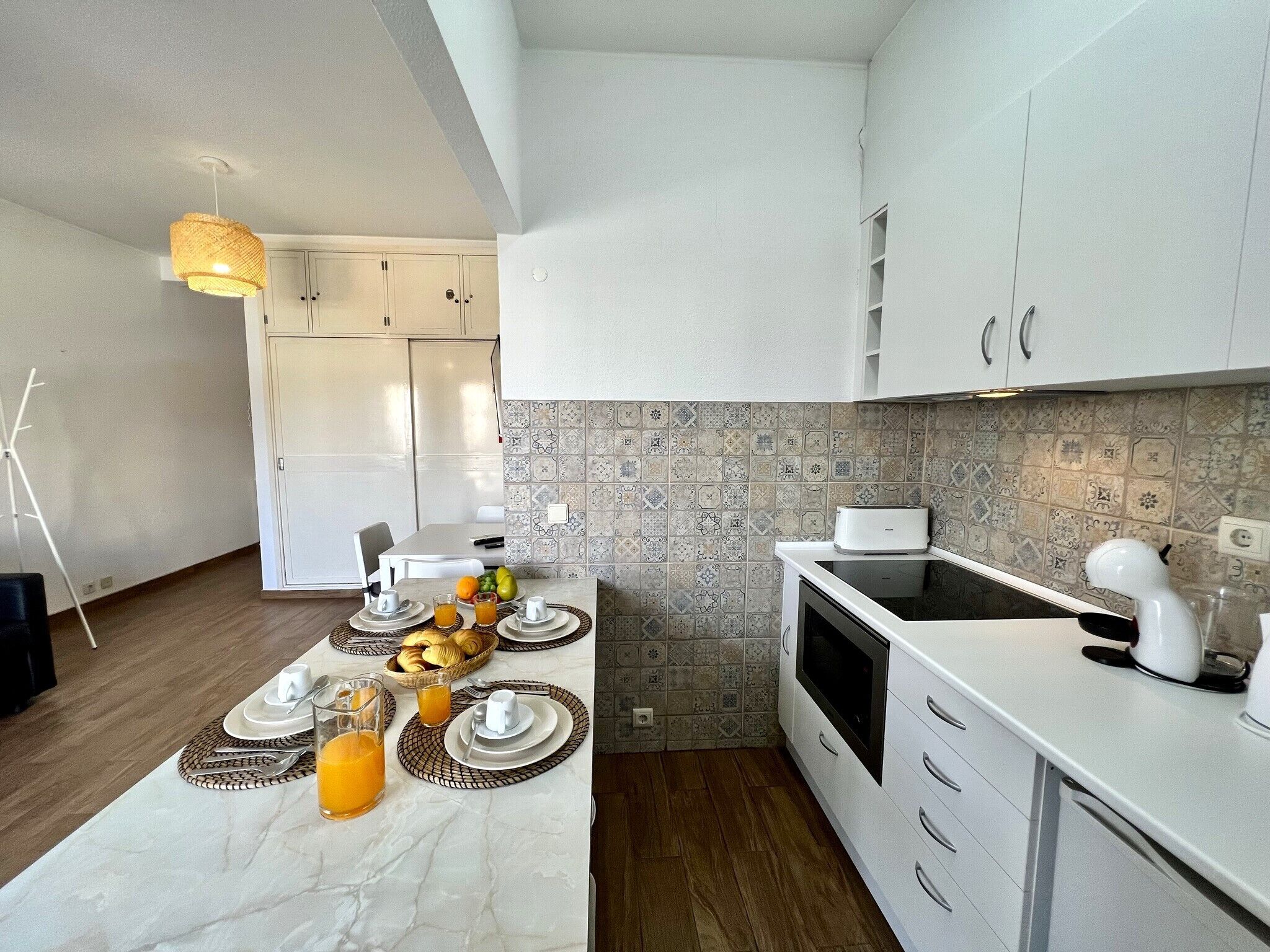 Een moderne keuken met witte kastjes, een betegelde spatwand, en een eethoek compleet met borden, glazen en een schaal fruit.