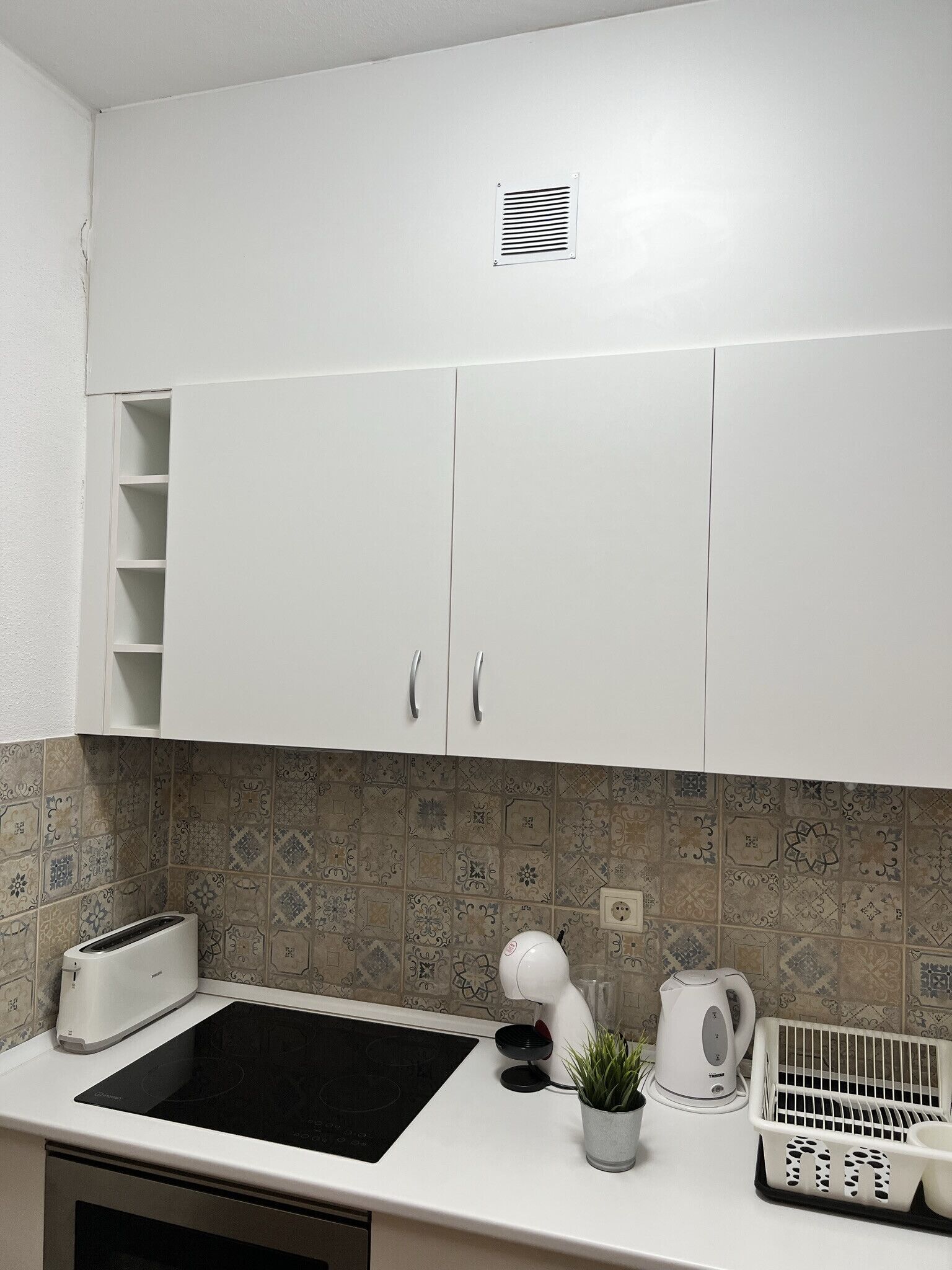 Una cocina con armarios blancos, una placa de inducción negra, una tetera, un tostador y una planta en la encimera.
