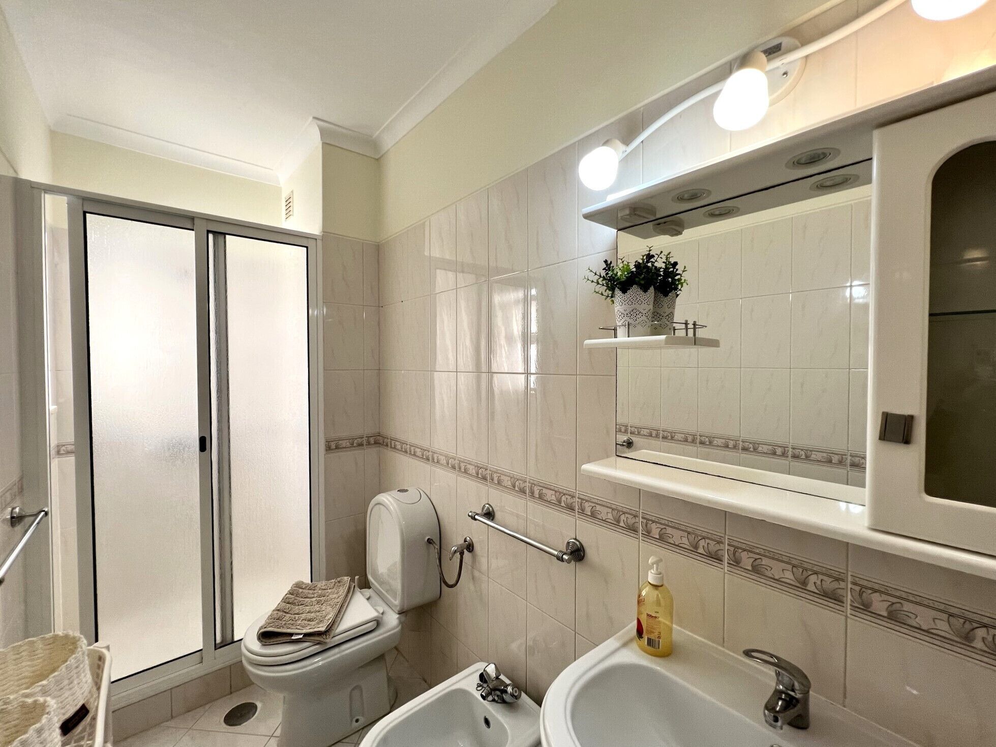 A bathroom with a toilet, bidet, and sink. There is a shower door, a towel, and a soap dispenser.