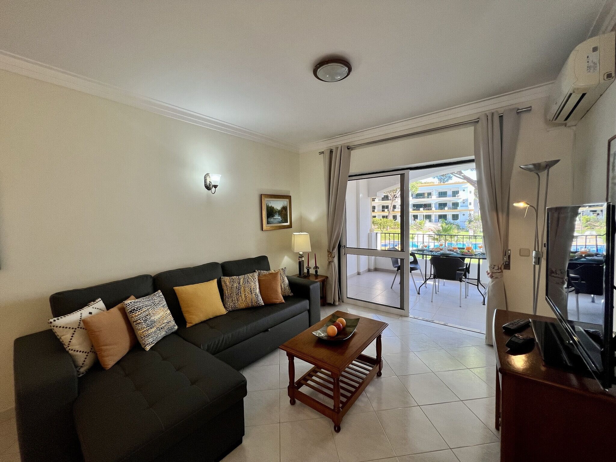 A living room with a sofa, a coffee table, a TV, and a balcony with outdoor furniture.