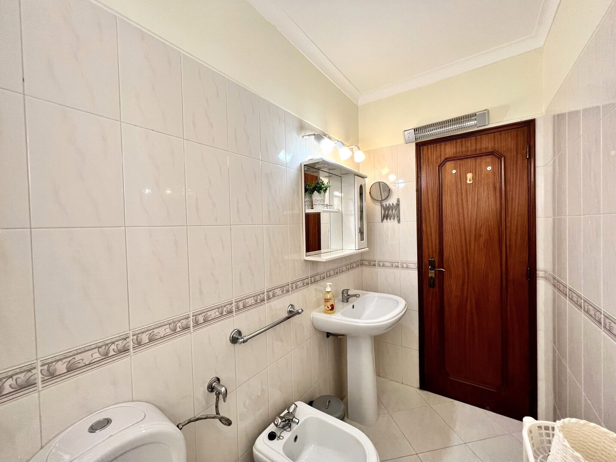 Ein Badezimmer mit einem Standwaschbecken, einer Toilette, einem Bidet, einer Holztür und einem Spiegel über dem Waschbecken.