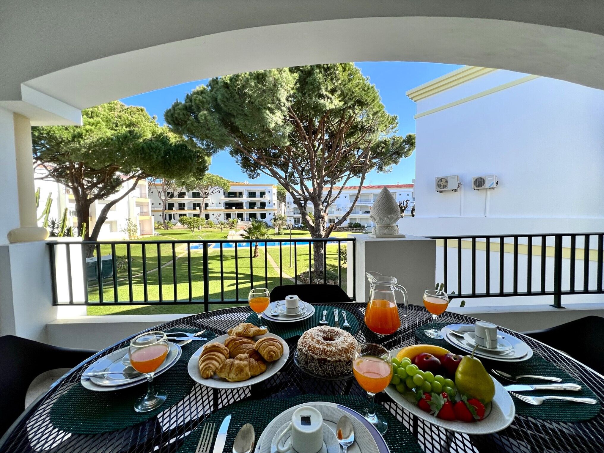 A balcony table set with breakfast items, a view of a pool, and a building in the background.