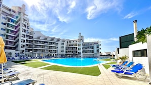 A swimming pool with blue lounge chairs and a grassy area, surrounded by multi-story buildings with balconies. - Albufeira SKY Light With Pool by Homing (Albufeira)