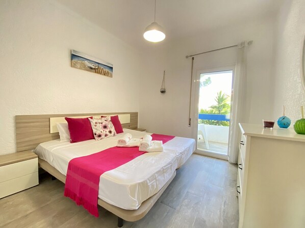 A hotel room with a bed, a nightstand, a desk with a chair, a window with a view of the outdoors, and a ceiling light.