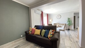 A living room with a dark brown sofa, colorful cushions, a dining area with a table and chairs, a clock on the wall, and a potted plant. - Quarteira Ocean Breeze 2 by Homing (Loulé)