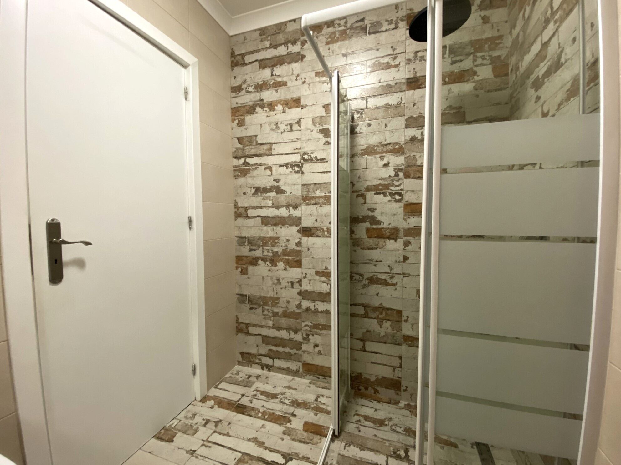 A shower with a glass door, a white door, and a brick-patterned wall.
