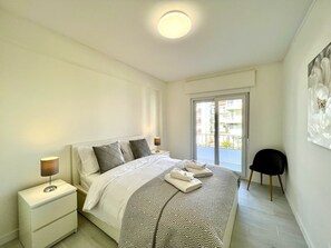 Quarto moderno com cama, mesas de cabeceira, uma cadeira e vista para uma varanda.