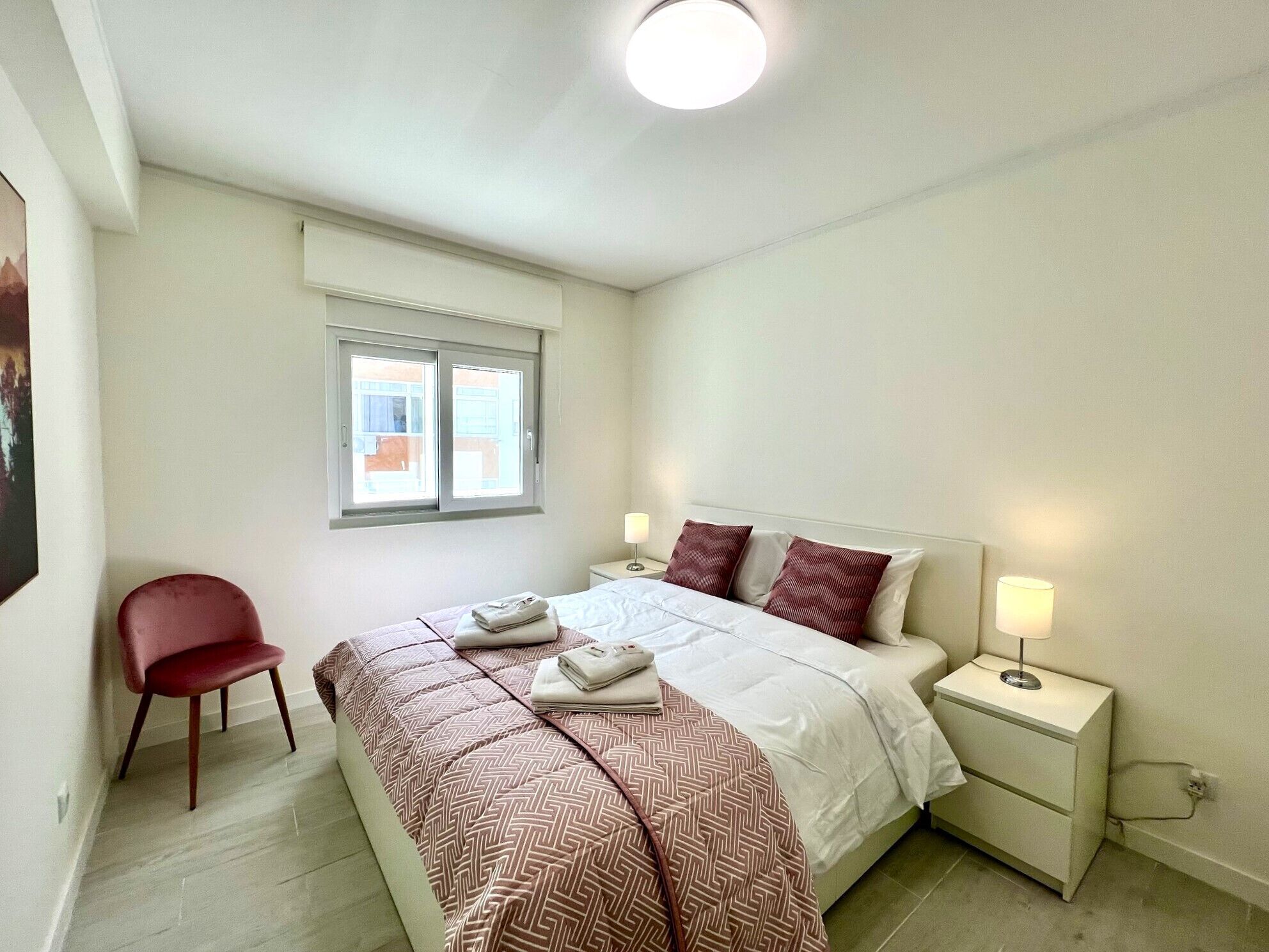 Un dormitorio ordenado con cama, mesitas de noche, una silla y un ventanal.