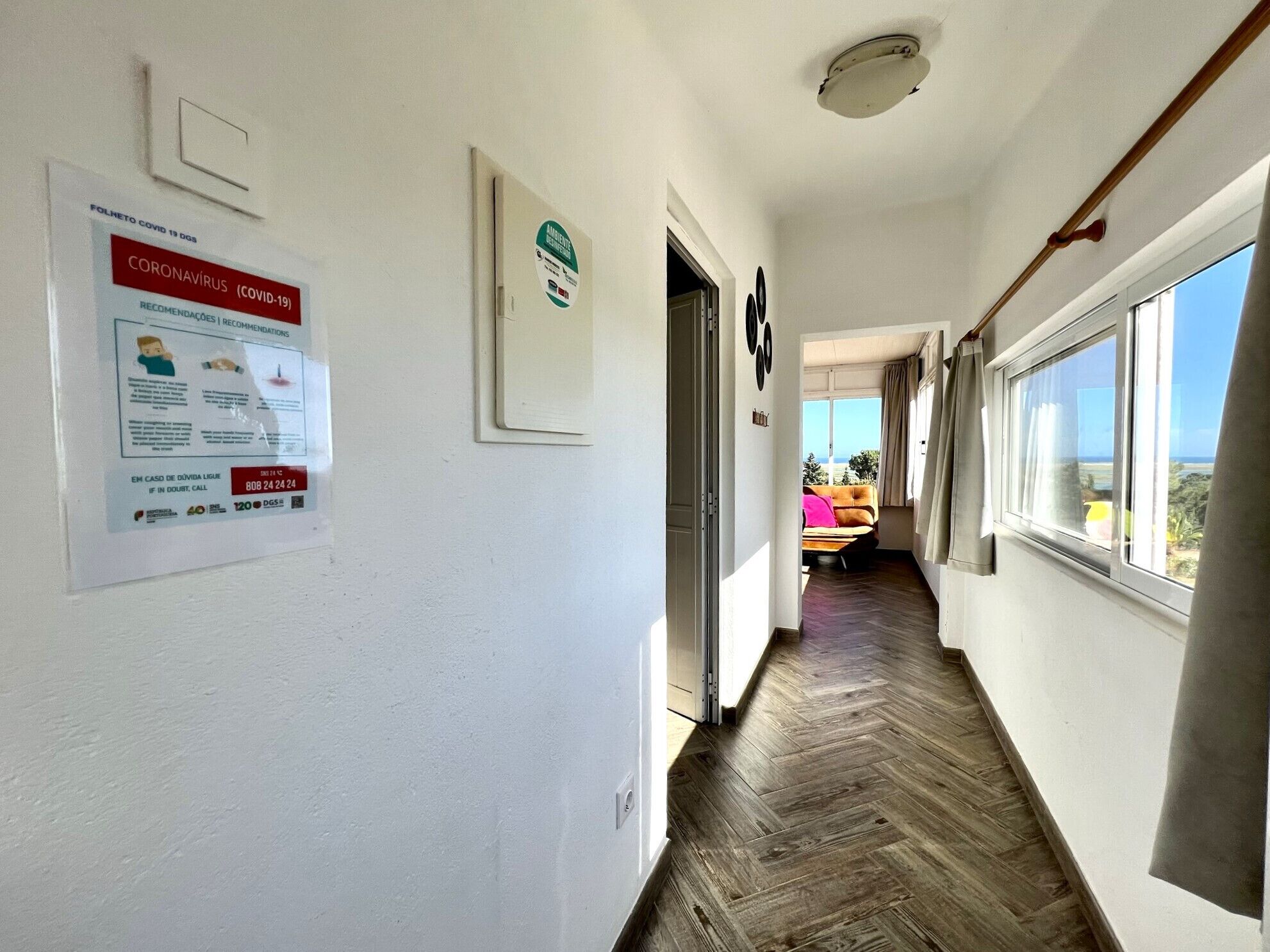 A hallway with a wooden floor, a bed with a pink pillow, and a window with a view of the ocean.