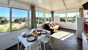 A sunlit room with a dining table set for breakfast, a sofa with cushions, and large windows offering a view of the outdoors. - Tavira Vila Formosa 6 With Pool (Tavira)
