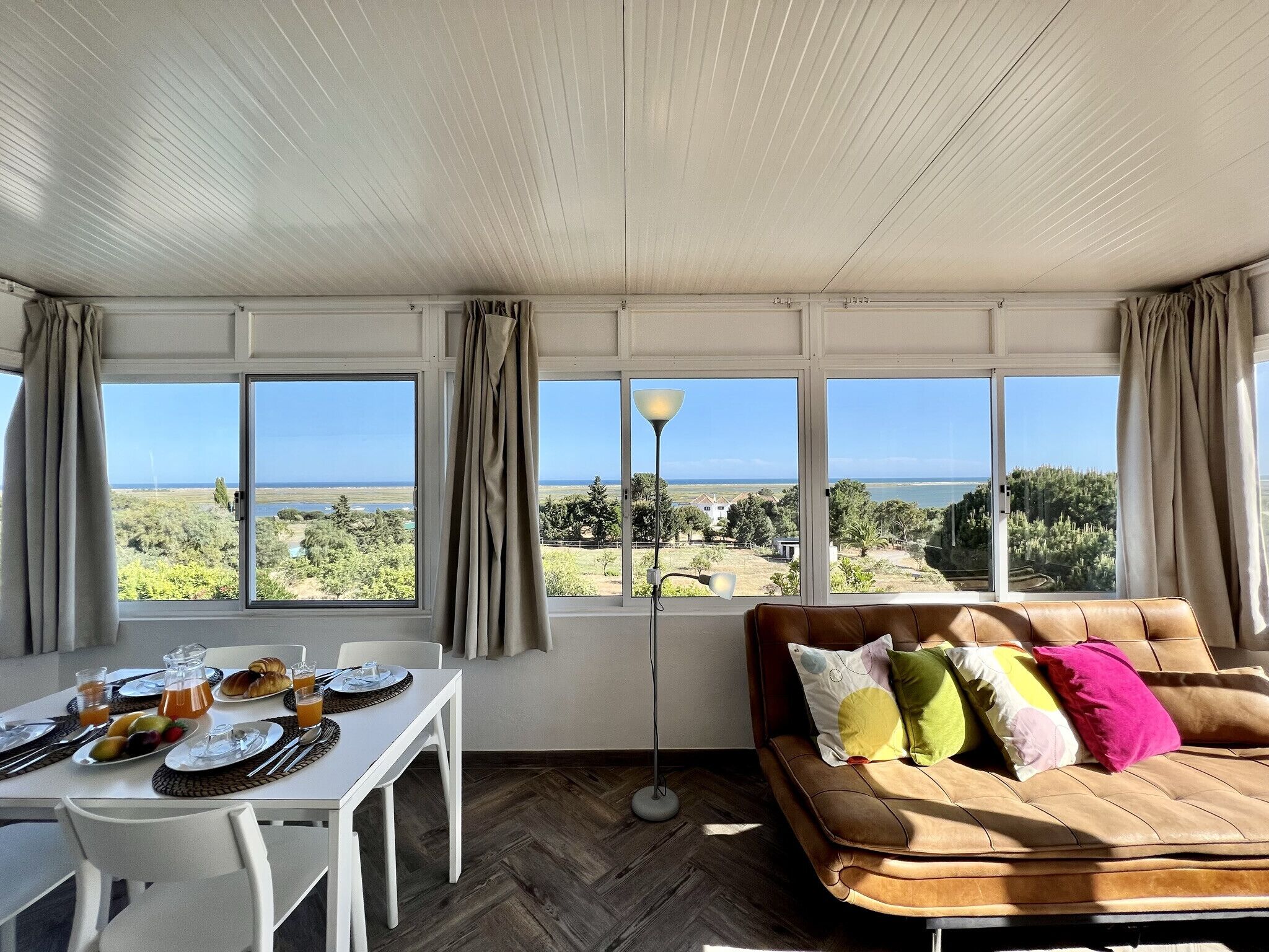 A room with a dining table set for breakfast, a sofa with colorful pillows, and large windows offering a view of the landscape.