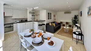 Una cocina moderna con armarios blancos, un comedor con una mesa blanca dispuesta para el desayuno y una sala de estar con un sofá oscuro y una planta.
