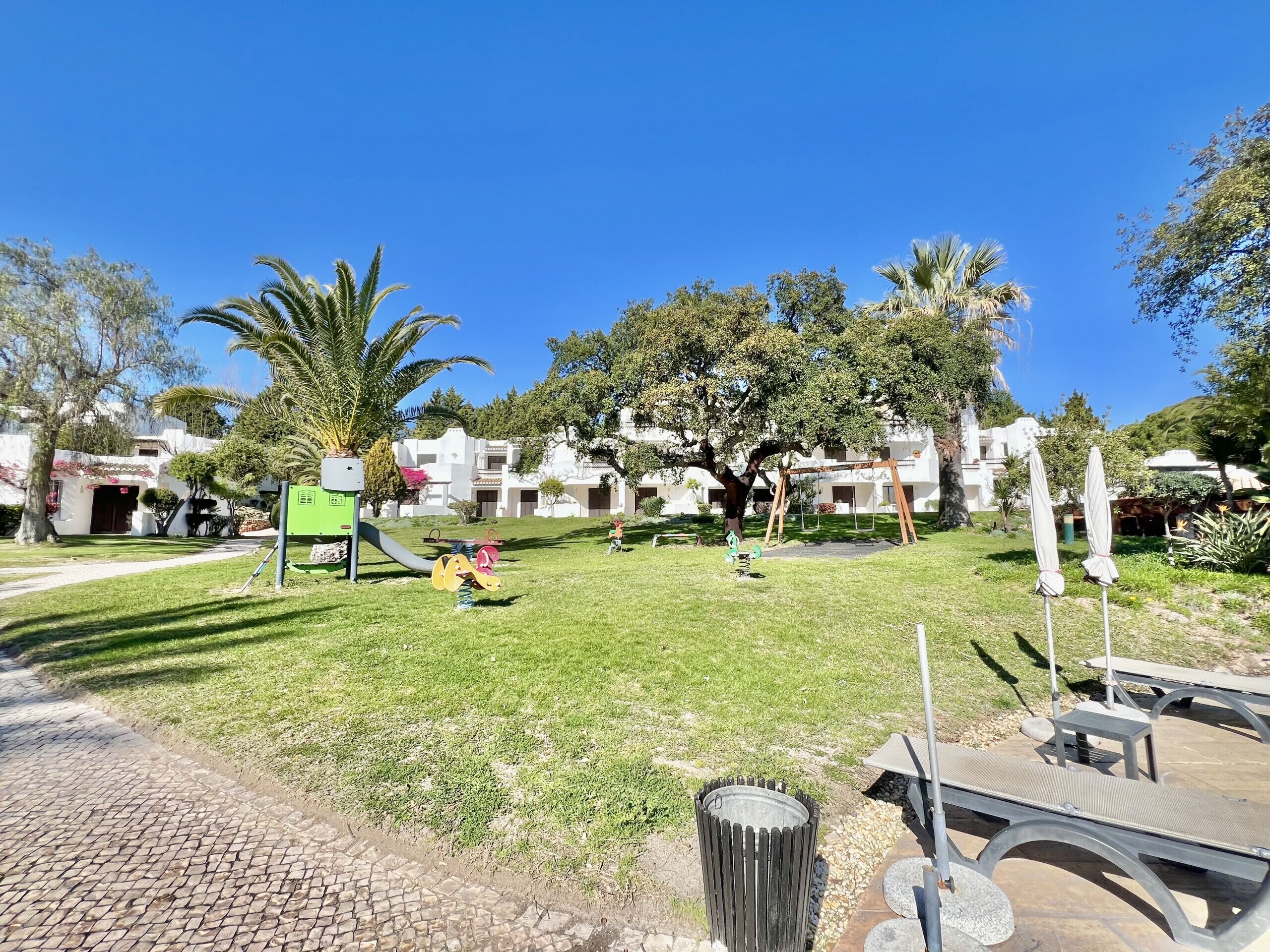 A playground with a slide and swings, surrounded by palm trees and a building with a white facade.