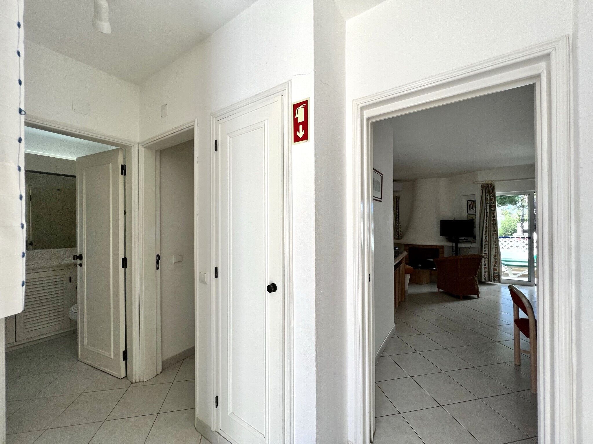 A hallway with white doors, a fire exit sign, and a dining area with a TV and chairs.