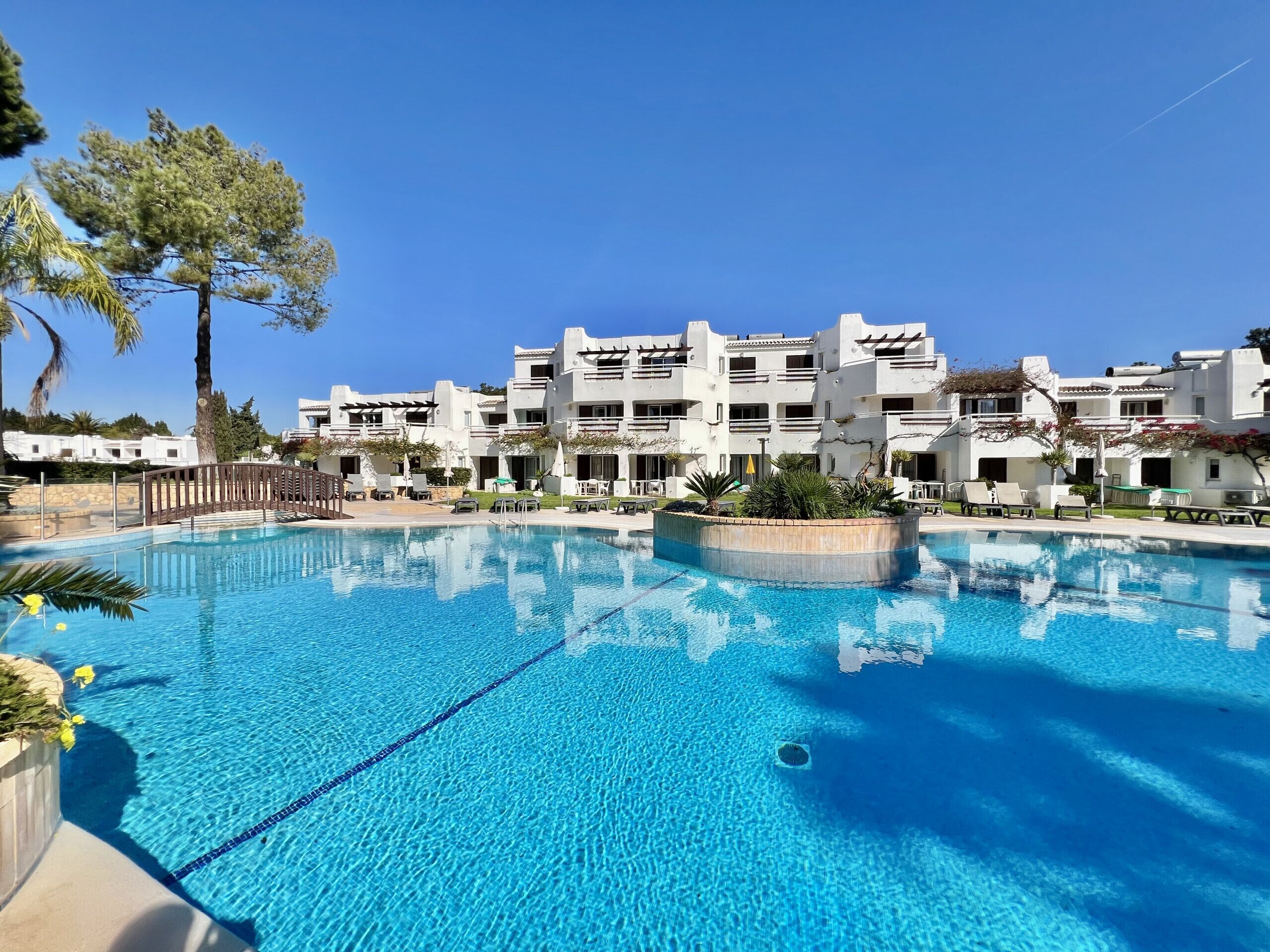 A large swimming pool with clear blue water, surrounded by white buildings with balconies and outdoor seating areas.