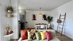 Una sala de estar moderna con un sofá, cojines de colores, una zona de comedor con mesa y sillas, una cocina al fondo, y cestas decorativas y plantas.