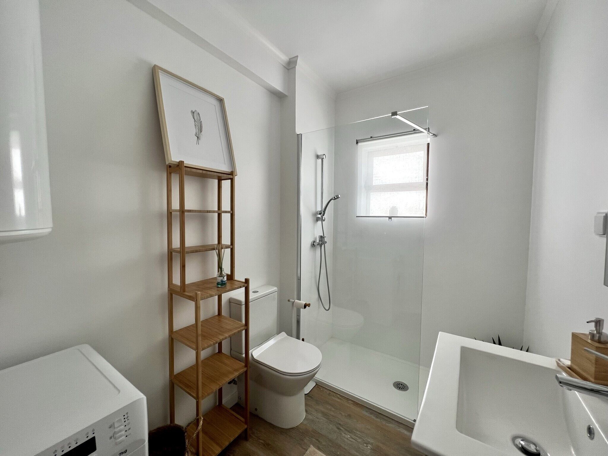 Et moderne badeværelse med en glasbrusekabine, et hvidt toilet, en træhylde med et indrammet billede og en hvid vask.