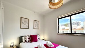 Una habitación de hotel con cama, mesita de noche, lámpara, dos cuadros enmarcados y ventana con vistas a la ciudad.