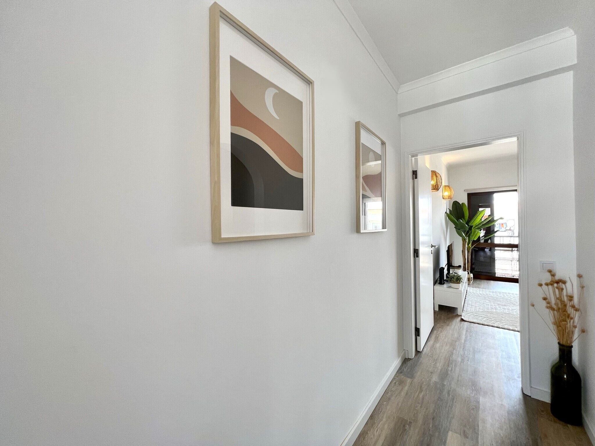 A hallway with white walls and wooden flooring, leading to a room with a large window and a plant.