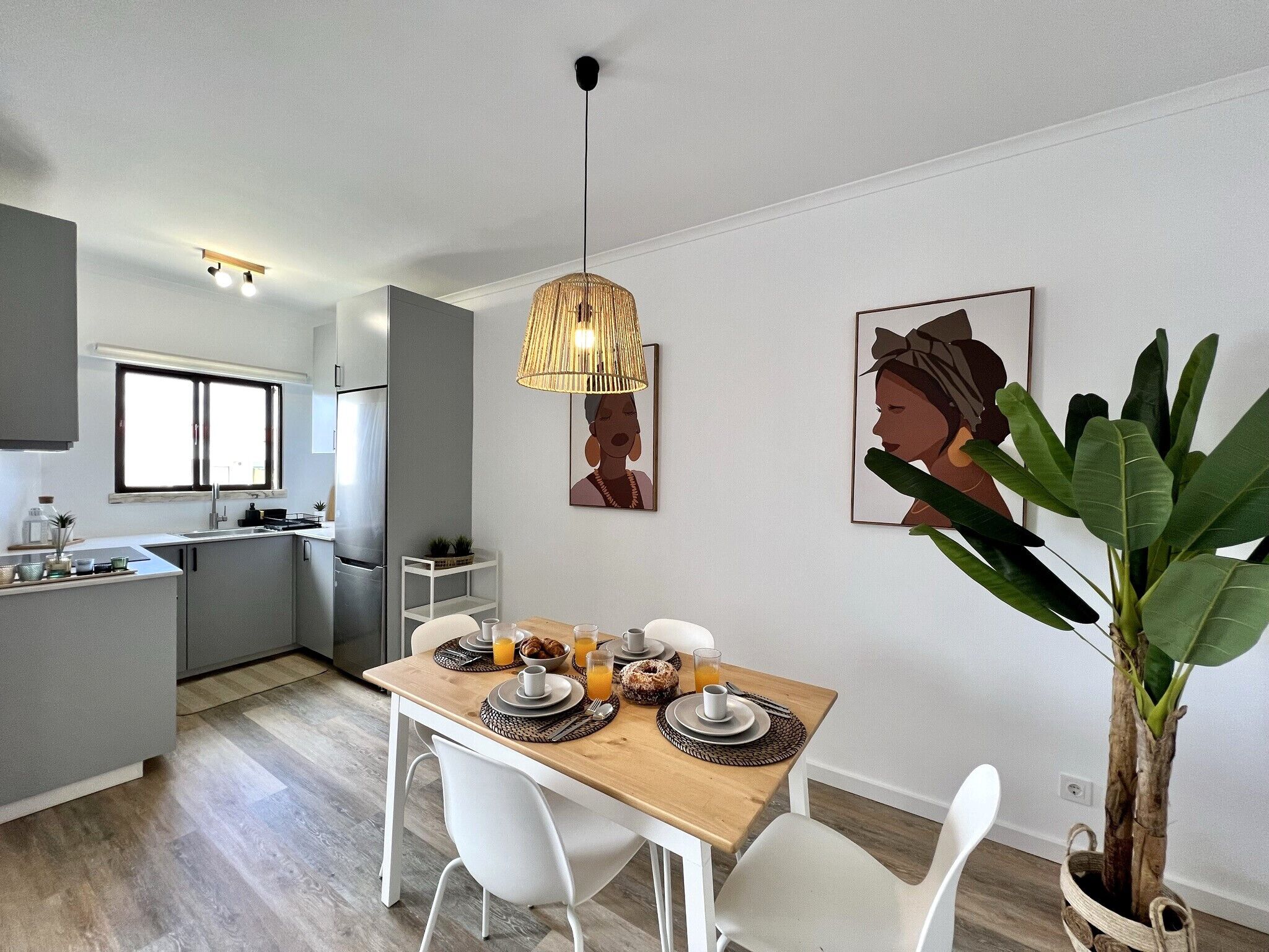A modern kitchen with a dining area, featuring a wooden table set for breakfast, a hanging light fixture, and a potted plant.
