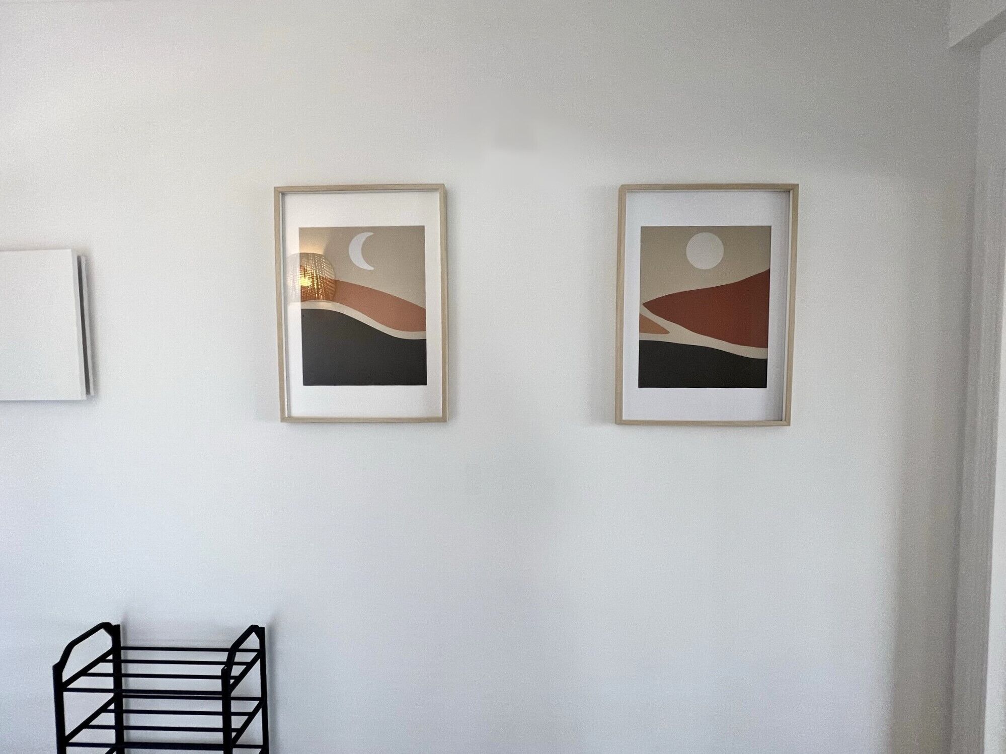Two framed abstract paintings on a white wall, a black metal rack, and a white object on the left.
