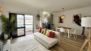Una sala de estar moderna con un sofá blanco, un comedor con mesa y sillas blancas, y una cocina al fondo.